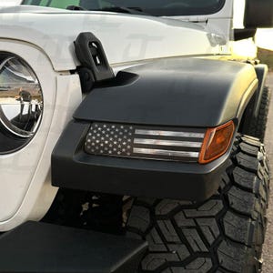 May include: A white Jeep Wrangler with a black and white American flag decal on the front fender. The decal is located above the front turn signal.
