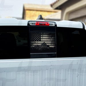 May include: A white pickup truck with a black and white American flag and mountain scene decal on the rear window.