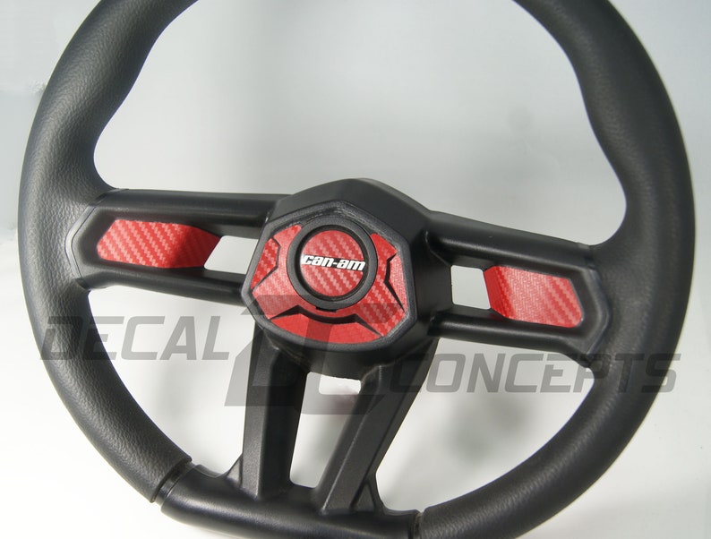 K&ouml;nnte beinhalten: Schwarzes Lenkrad mit roten Kohlefaser-Akzenten und dem "Can-Am"-Logo. Das Lenkrad hat einen strukturierten Griff und eine zentrale Nabe mit geometrischem Design. Der Text "DECAL CONCEPTS" ist ebenfalls zu sehen.