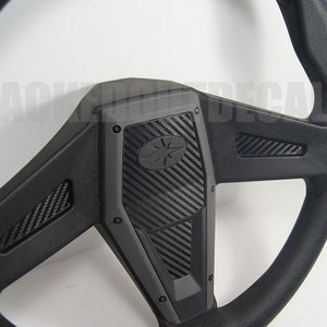 May include: Black steering wheel with a carbon fiber design. The steering wheel has a black leather rim and a black plastic center with a carbon fiber pattern.