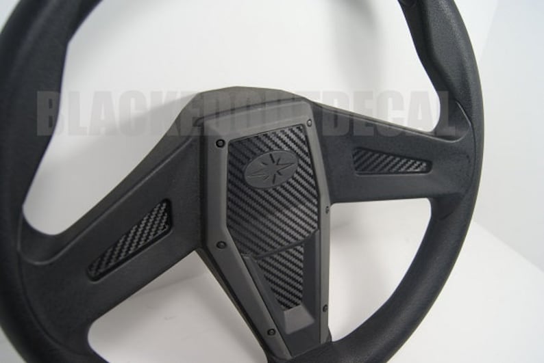May include: Black steering wheel with a carbon fiber design. The steering wheel has a black leather rim and a black plastic center with a carbon fiber pattern.