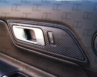 May include: Close-up of a car door interior with a black carbon fiber trim panel. The panel features a silver door handle and a black button with a lock icon.