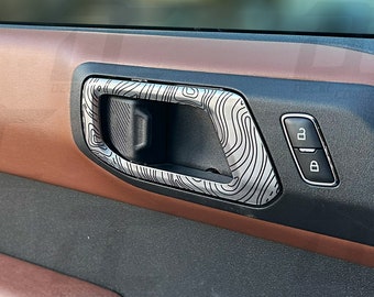 Peut inclure: Gros plan sur l'intérieur d'une portière de voiture avec une poignée de porte à motif bois et un bouton noir avec la lettre "B" dessus.