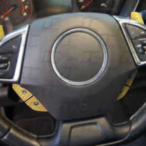 May include: A black leather steering wheel with yellow carbon fiber accents. The steering wheel has buttons on the left side and a plus and minus button on the right side.