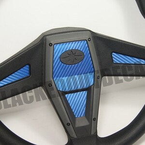 May include: Black steering wheel with blue carbon fiber accents. The steering wheel has a star-shaped logo in the center.