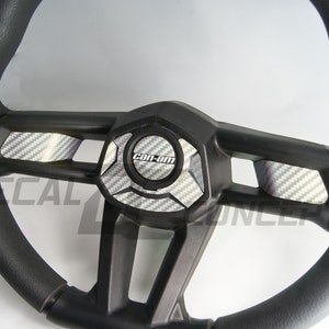 K&ouml;nnte beinhalten: Schwarzes Lenkrad mit strukturierter Grifffl&auml;che. Die Mitte zeigt eine schwarze Platte mit dem "Can-Am"-Logo und Akzenten im Carbonfaser-Stil. Das Lenkrad hat ein sportliches Design, geeignet f&uuml;r Gel&auml;ndefahrzeuge.