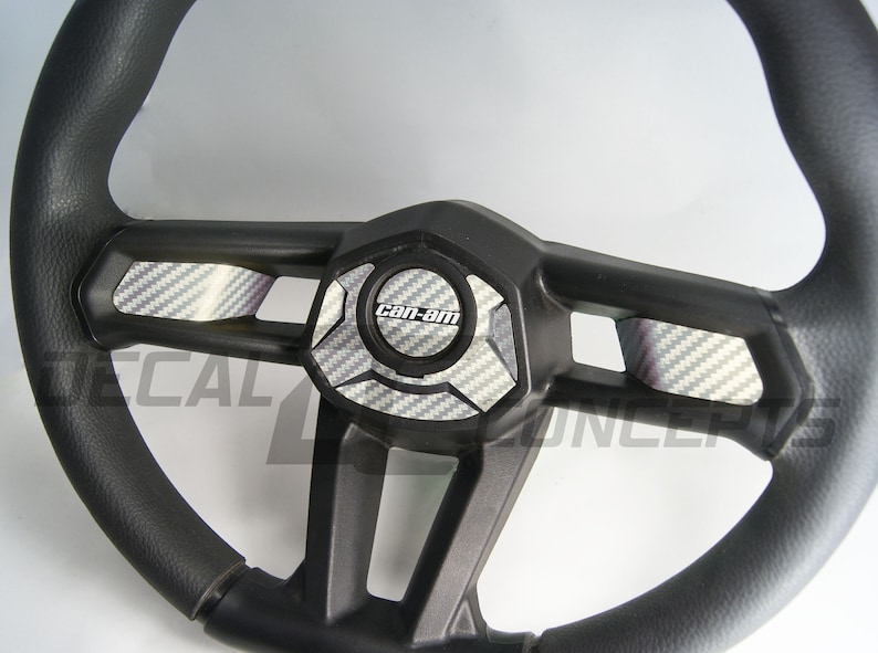 K&ouml;nnte beinhalten: Schwarzes Lenkrad mit strukturierter Grifffl&auml;che. Die Mitte zeigt eine schwarze Platte mit dem "Can-Am"-Logo und Akzenten im Carbonfaser-Stil. Das Lenkrad hat ein sportliches Design, geeignet f&uuml;r Gel&auml;ndefahrzeuge.