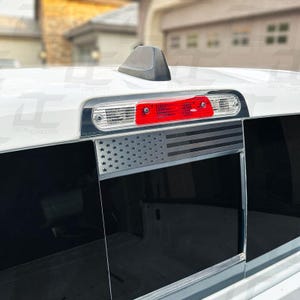 May include: A black and white American flag decal on the back window of a white pickup truck. The decal is placed above the taillight and has a silver finish.
