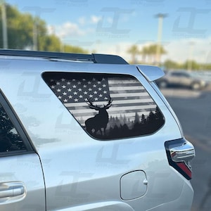 May include: A silver SUV with a rear window decal featuring a black and white American flag with a silhouette of a deer in a forest scene.