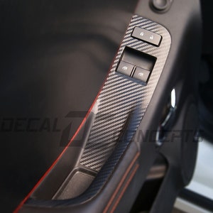 May include: Close-up of a car's interior door panel with a carbon fiber pattern. The panel features window controls and a lock button. The panel is accented with a red stitching detail.