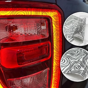 May include: A vehicle taillight assembly featuring a red and orange lens with a black topographic map pattern overlay. Two circular insets display the adhesive vinyl film roll, showcasing the black wavy line design on a clear background.