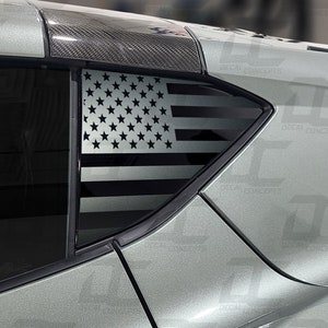 May include: A silver and black American flag decal on a car window. The flag is in the style of a distressed or weathered look.