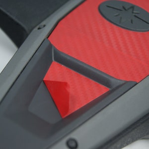 May include: Close-up of a black and red steering wheel with a carbon fiber pattern. The steering wheel has a black center with a red accent and a black star logo.
