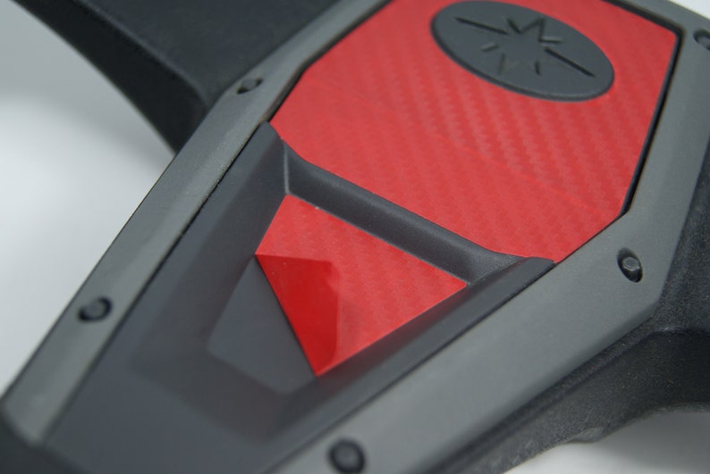 May include: Close-up of a black and red steering wheel with a carbon fiber pattern. The steering wheel has a black center with a red accent and a black star logo.