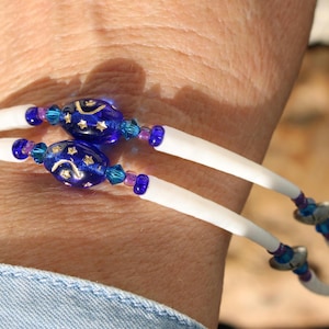 May include: A double-strand bracelet with white bands and blue star-patterned beads. The bracelet features small blue and purple accent beads. The jewelry is worn on a wrist, with a blue denim shirt visible.