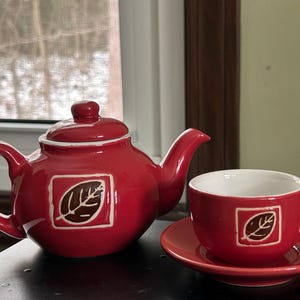 May include: A red ceramic teapot with a matching teacup and saucer set. The teapot and teacup feature a leaf design within a square frame. The set is placed on a dark surface.