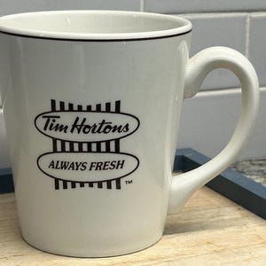 May include: White ceramic coffee mug with a black rim and handle. The mug features the Tim Hortons logo and the words "ALWAYS FRESH" in black. The mug is on a wooden surface.