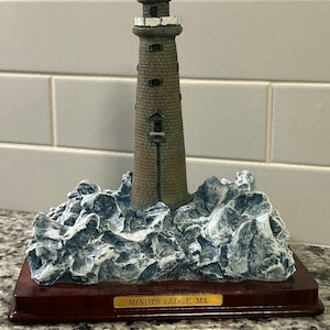 May include: A miniature lighthouse figurine of Minots Ledge, MA, featuring a tall, textured brown stone tower with a lit yellow lantern room, set upon a base of sculpted blue and white crashing waves on a dark brown rectangular wooden plinth.