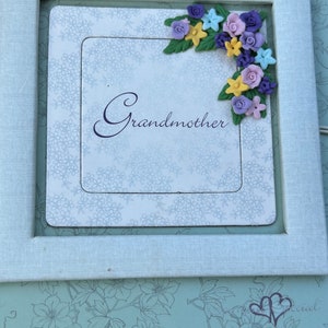 May include: A white frame with a light blue background and a floral design. The frame has a white mat with the word "Grandmother" written in black cursive. The frame is decorated with a cluster of colorful flowers in the upper right corner.