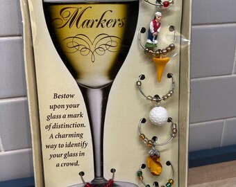 May include: A set of six wine glass markers in a box. The markers are themed around golf and feature a golfer, a golf ball, a golf tee, a golf glove, and two golf flags. The box has the text "Boston Warehouse® Wine Markers" on the front.