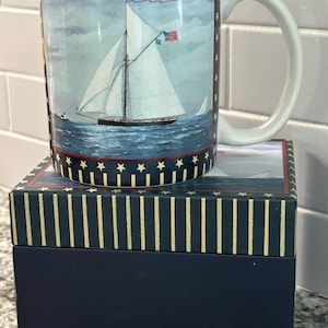 May include: A ceramic mug with a nautical theme, featuring a sailboat on the ocean. The mug has a white handle and is decorated with a blue and red border with stars. It sits on a matching navy blue box with vertical stripes.