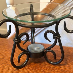 May include: A dark brown metal oil warmer featuring a circular frame with decorative scrollwork legs and a clear glass bowl resting on top. A small, round, light blue tea light candle sits in the center base of the metal structure.