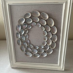 Puede incluir: Obra de arte enmarcada con un diseño circular de conchas de abulón iridiscentes. Las conchas están dispuestas sobre un fondo de tela gris dentro de un marco de madera blanco, creando una estética costera.