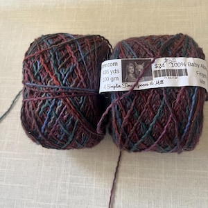 May include: Two skeins of multi-colored yarn. The yarn is a mix of burgundy, teal, and blue. Each skein has a label that reads "100% Baby Alpaca" and "436 yds, 100 gm". The yarn is from "A Simpler Time pacas & Mill".