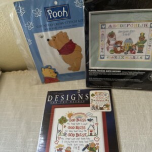Puede incluir: Tres kits de punto de cruz contado en envases de plástico. Un kit presenta a Winnie the Pooh, otro es un registro de nacimiento Dimensions Playful Teddies y el tercero es un kit Designs for the Needle con el texto God Bless y figuras.