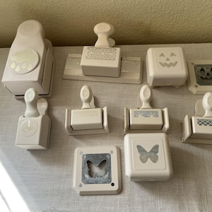 May include: A collection of off-white paper punches with various designs, including a cloud with raindrops, a butterfly, and a jack-o'-lantern. The punches are arranged on a light-colored surface.