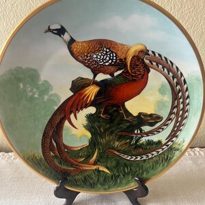 May include: A round decorative porcelain plate featuring a detailed illustration of two pheasants perched on a mossy log. The birds display vibrant orange, red, brown, and black plumage with long, patterned tail feathers against a soft sky background.