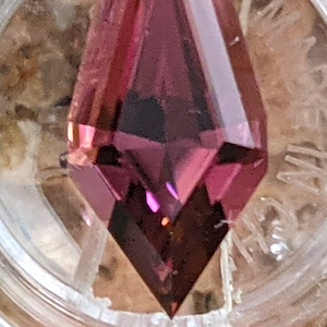 May include: A deep purple gemstone with a faceted, pointed shape. The gemstone is cut in a unique, elongated style.