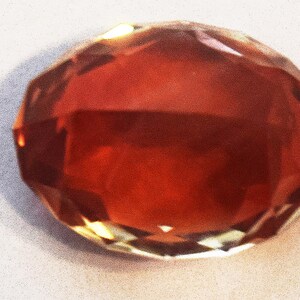 May include: A faceted, red-brown gemstone with a high shine. The gemstone is cut into a round shape with many facets.