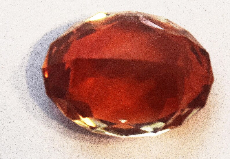May include: A faceted, red-brown gemstone with a high shine. The gemstone is cut into a round shape with many facets.