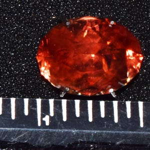 May include: A large, oval-shaped, red gemstone with a faceted cut. The gemstone is set on a black background.