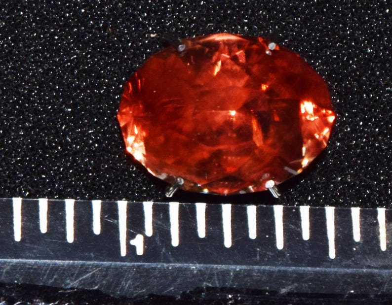 May include: A large, oval-shaped, red gemstone with a faceted cut. The gemstone is set on a black background.