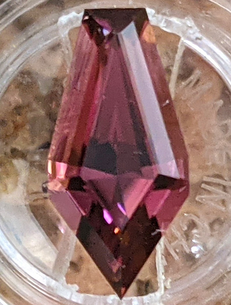 May include: A deep purple gemstone with a faceted, pointed shape. The gemstone is cut in a unique, elongated style.