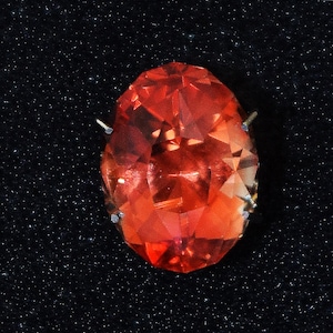 May include: A large, faceted, oval-shaped, red gemstone with a high polish. The gemstone is set in a silver or white metal setting with prongs on the side.