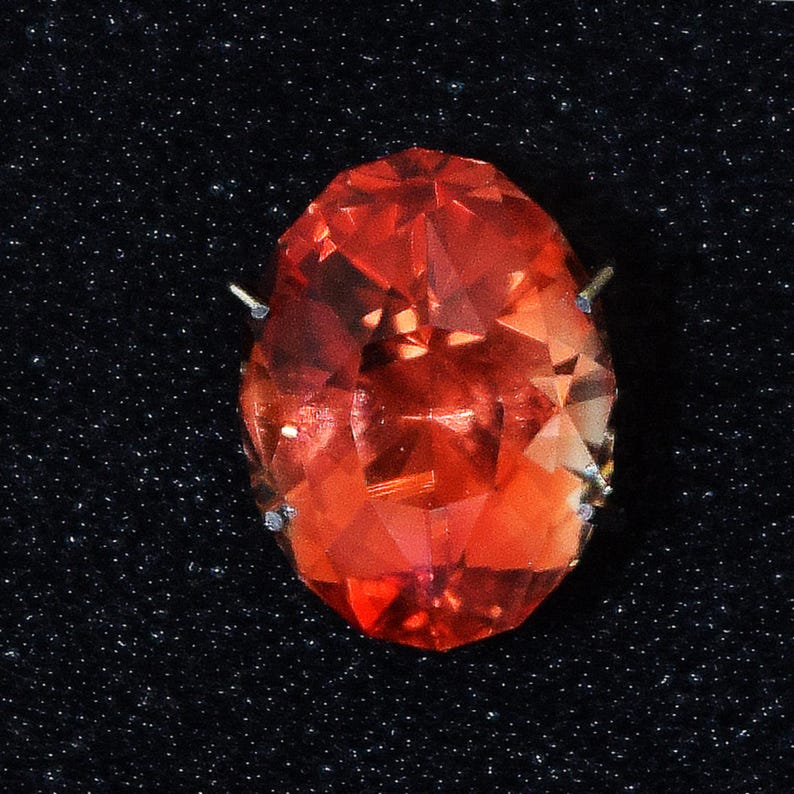May include: A large, faceted, oval-shaped, red gemstone with a high polish. The gemstone is set in a silver or white metal setting with prongs on the side.