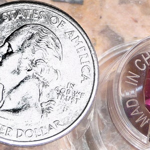 May include: A close-up of a purple gemstone, faceted and cut into a pointed shape, resting in a clear plastic container. The container is labeled "MADE IN CHINA". A US quarter is shown for scale.