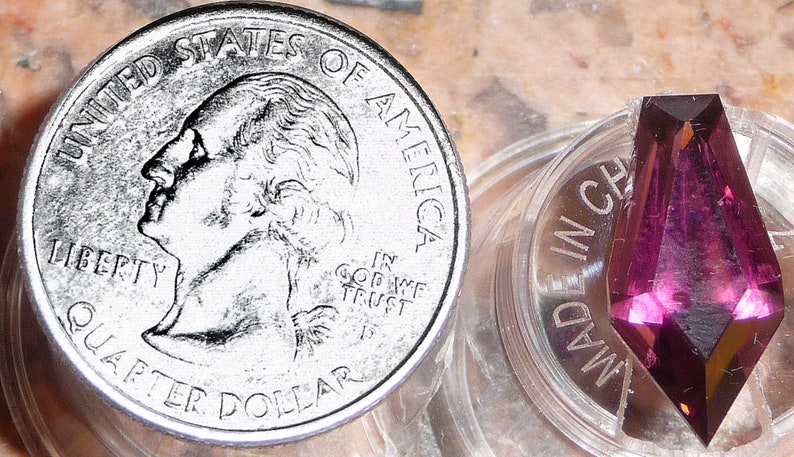 May include: A close-up of a purple gemstone, faceted and cut into a pointed shape, resting in a clear plastic container. The container is labeled "MADE IN CHINA". A US quarter is shown for scale.