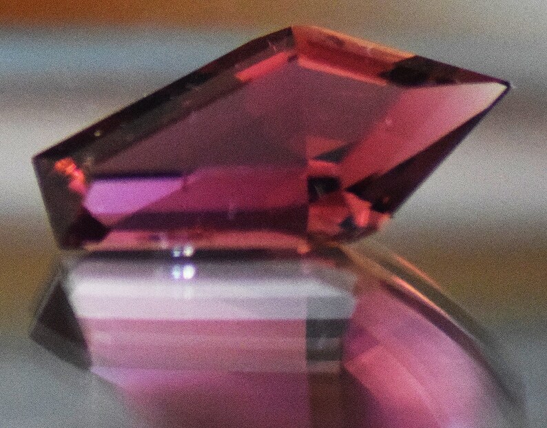 May include: A close-up of a pink gemstone with a faceted cut. The gemstone is set in a ring with a silver band.