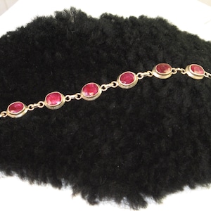 Op de afbeelding: Een zilveren armband met ovale rode edelstenen. De armband heeft een sluiting.