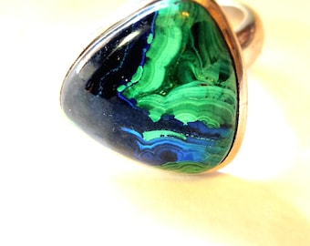 Könnte beinhalten: Ein Silberring mit einem dreieckigen Malachitstein. Der Stein zeigt ein wirbelndes Muster aus grünen, blauen und schwarzen Farbtönen. Der Ring hat ein poliertes Silberband und eine Lünettenfassung, die den Stein einfasst.