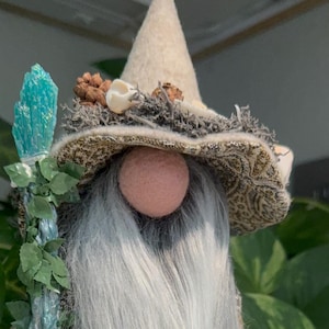 May include: A whimsical gnome figurine with a long white beard, wearing a patterned hat and coat. The gnome holds a blue crystal staff and has a seashell at its feet. A small charm hangs from its hand.