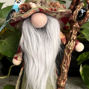May include: A green gnome with a long white beard wearing a green pointed hat with a red and gold patterned brim. The gnome is holding a brown wooden staff with a black orb on top. The gnome is decorated with a blue bird, pine cones, and other natural elements.