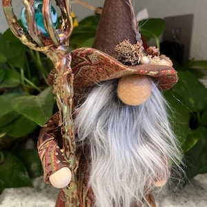 May include: A gnome wearing a brown hat with a feather and a gold and red patterned robe, holding a staff with a green glass orb on top.