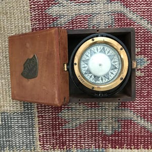 May include: A vintage compass in a wooden box. The compass has a brass face with a black needle and a white background. The box is made of dark wood and has a brass latch. The compass is labeled "YACHT "GRAY GHOST" 24.5 ft."
