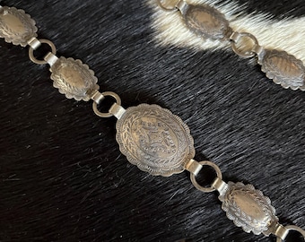 May include: A silver-toned metal chain belt composed of repeating oval-shaped links. Each link features a scalloped edge and intricate engraved patterns. The links are connected by small circular jump rings, creating a flexible, articulated design.
