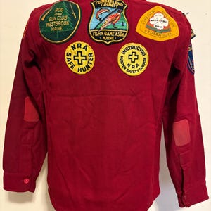 May include: A red long-sleeve shirt with multiple patches. Patches include "Rod and Gun Club Westbrook Maine", "Cumberland County Fish & Game Assn Maine", "NRA Safe Hunter", "Instructor NRA Safety Course", and "Virginia Wildlife Federation".